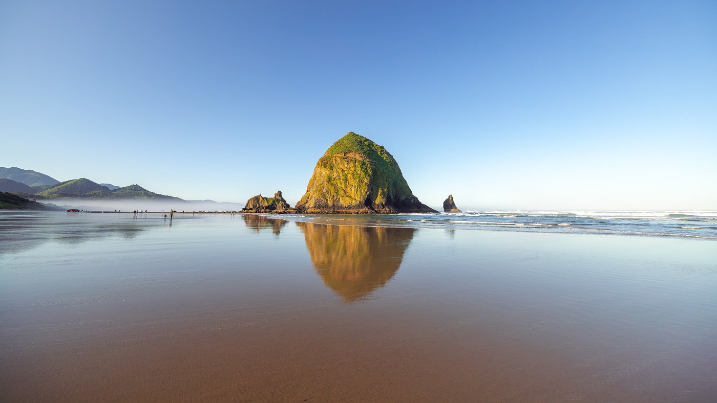 Haystack Rock reflected in the wet sand at Cannon Beach, Oregon, under a clear blue sky